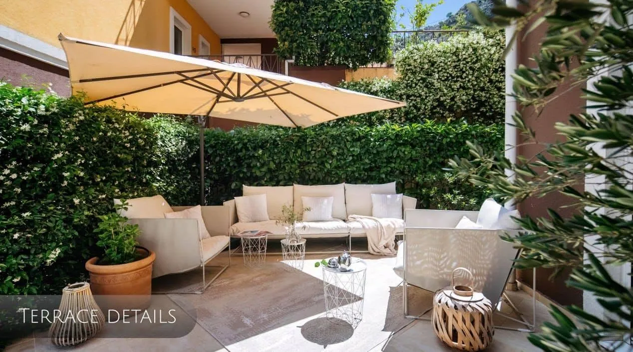 Cozy outdoor terrace with white furniture, a large umbrella, and green hedges surrounding the space.