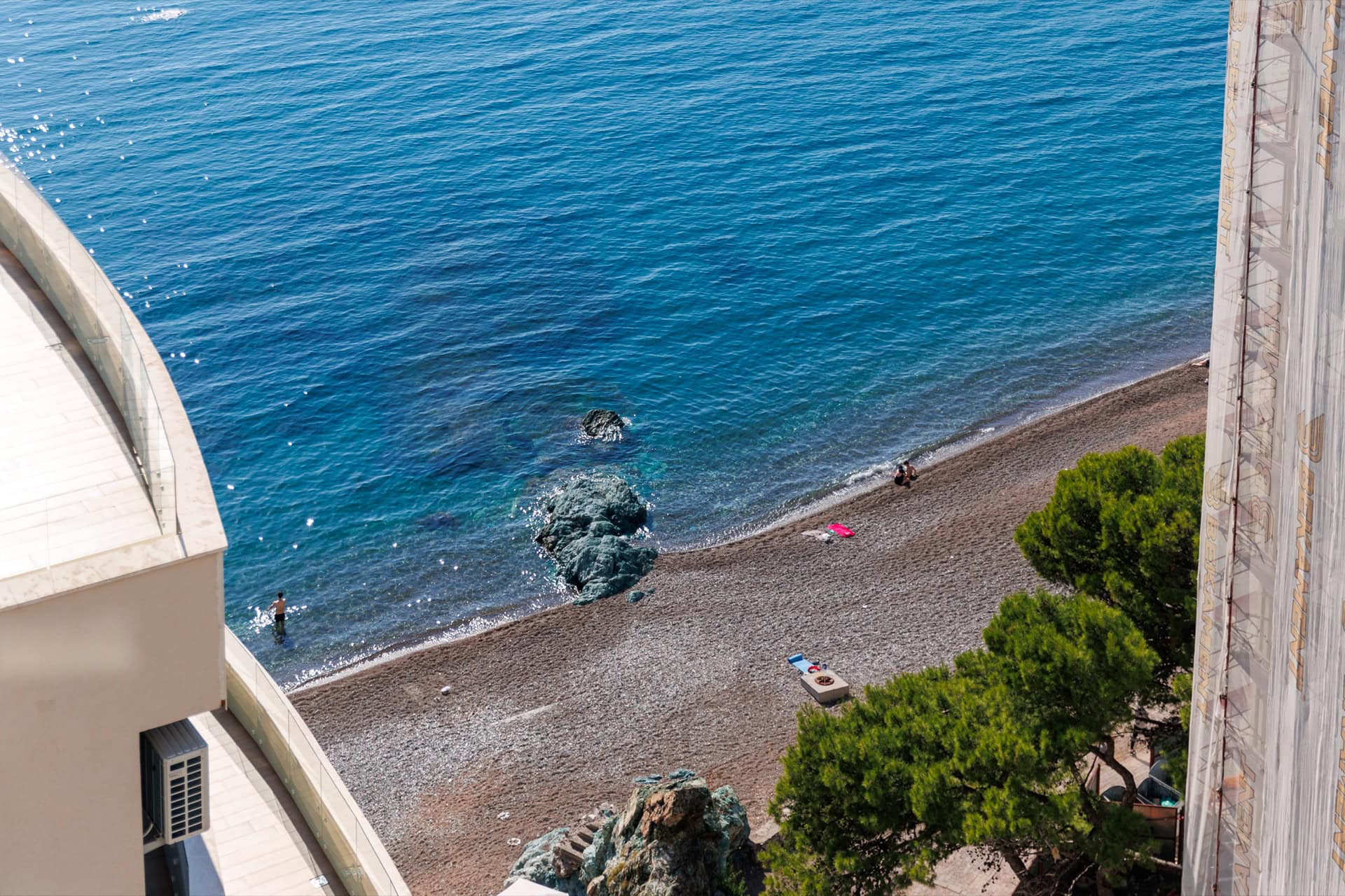 View from the apartment balcony in Bečići, Montenegro: a panorama of a private pebble beach and the azure Adriatic Sea.