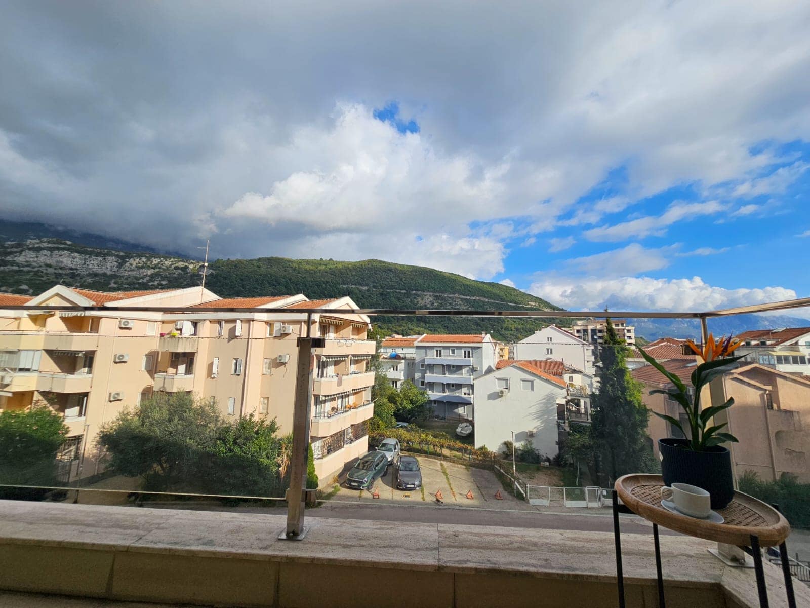 View from the apartment balcony in Budva, Montenegro: city panorama, mountains, and parking lot, with a table and flower in the foreground.