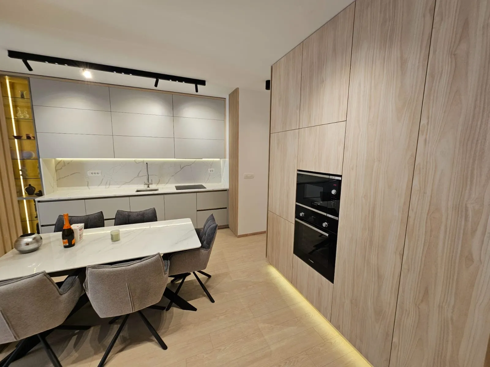 Modern kitchen with light wood cabinetry, white marble countertops, built-in oven and microwave, and a dining table with grey chairs.