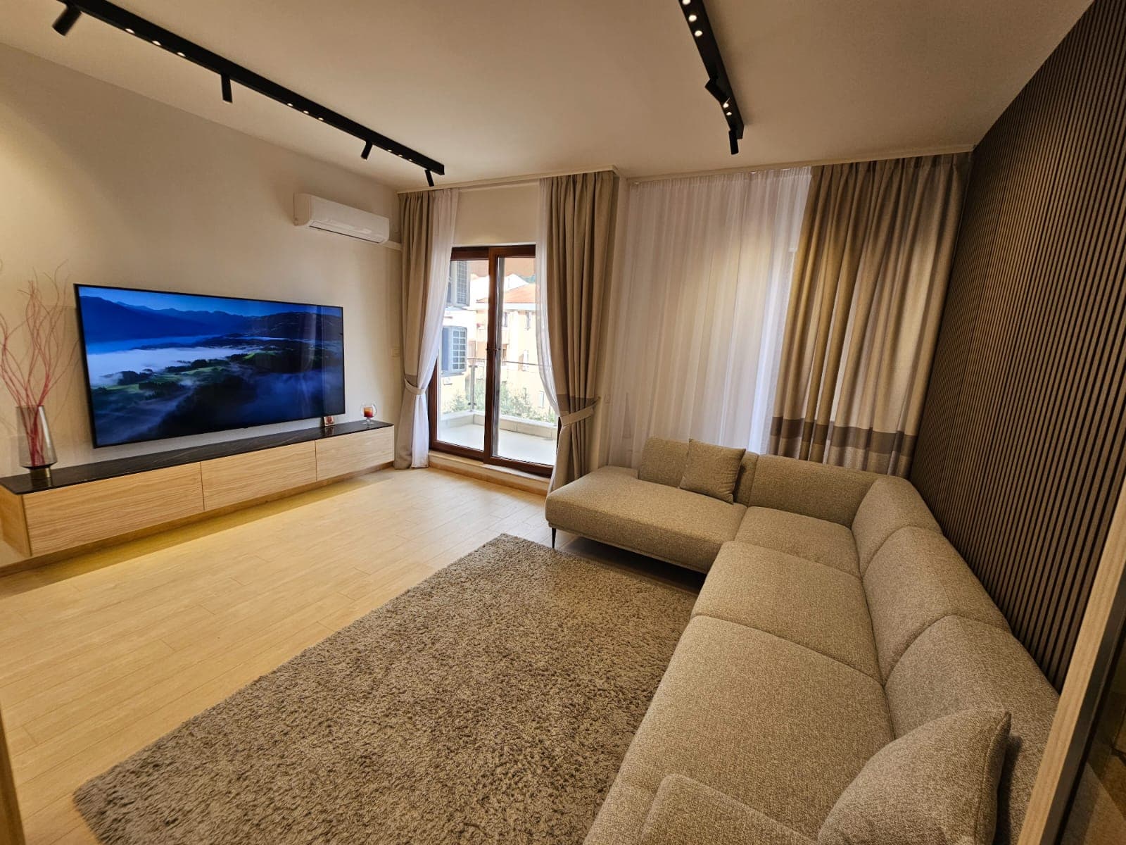 Modern living room in an apartment in Budva, Montenegro: corner sofa, TV area, slatted wall, and access to the balcony.