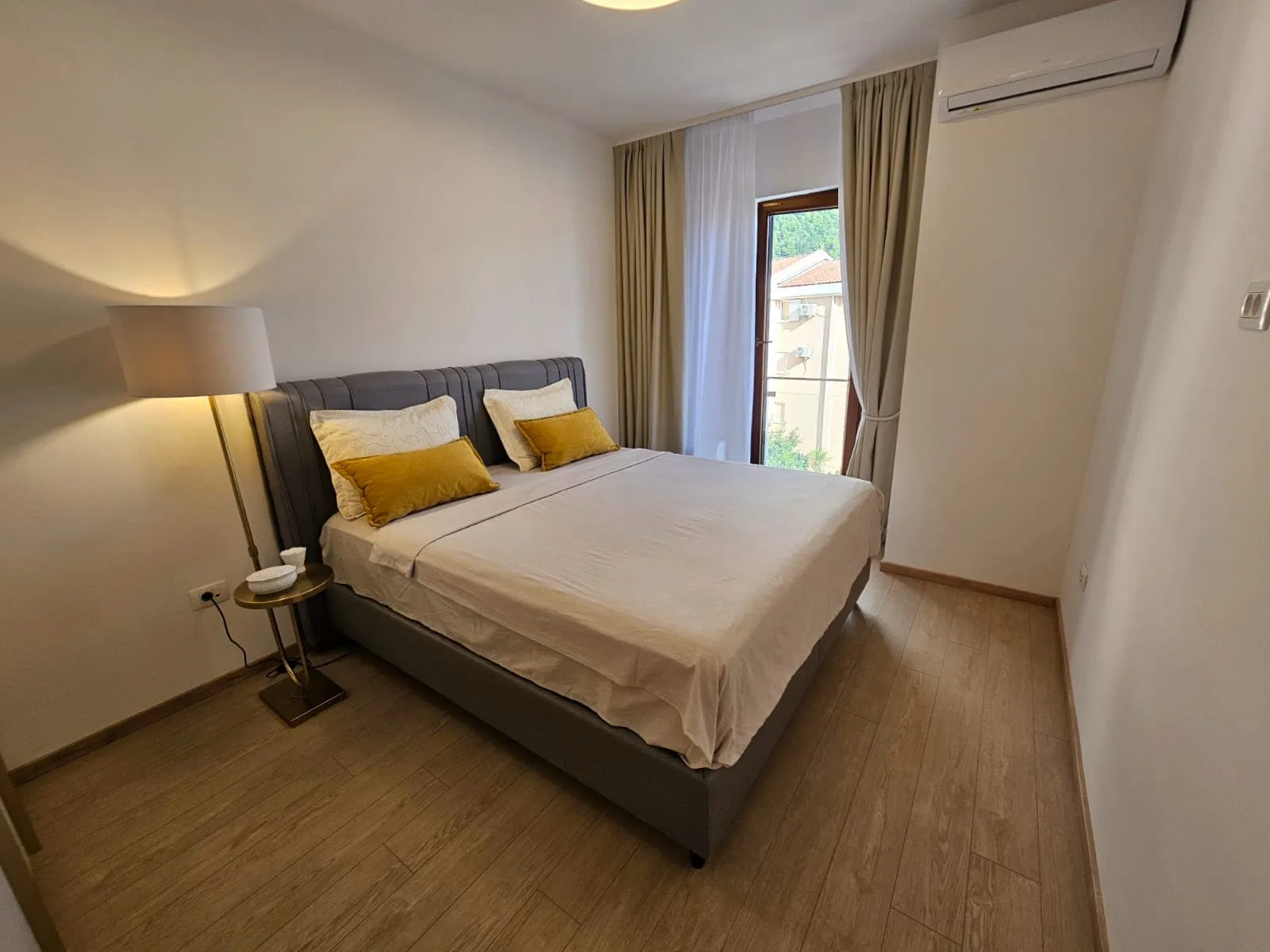 Bedroom featuring a double bed with a grey headboard, mustard pillows, a floor lamp, and wood flooring.