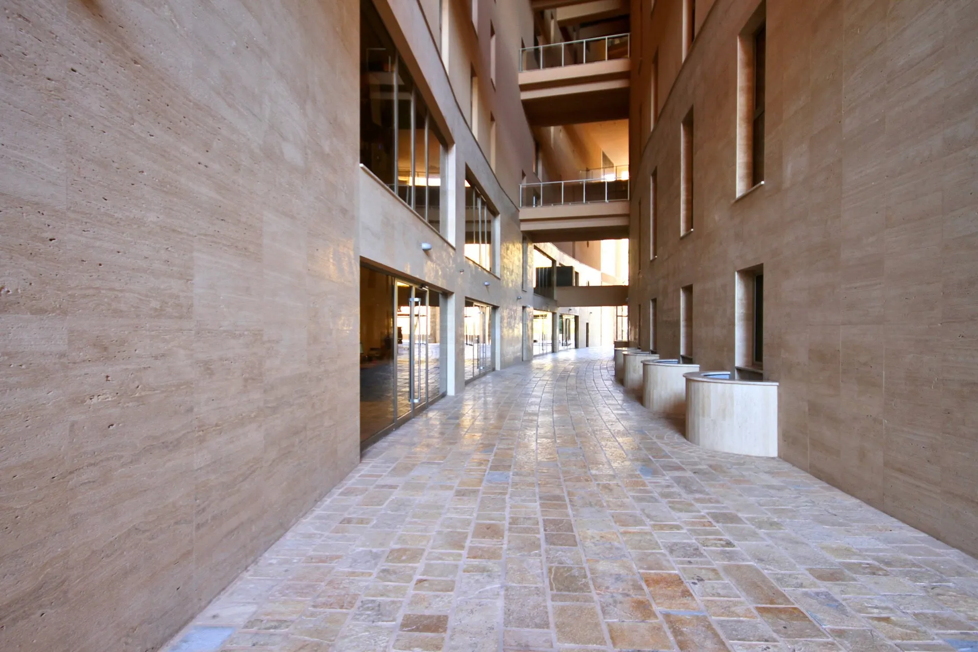 Long corridor with stone flooring and light stone walls, large windows, and balconies visible on upper levels.