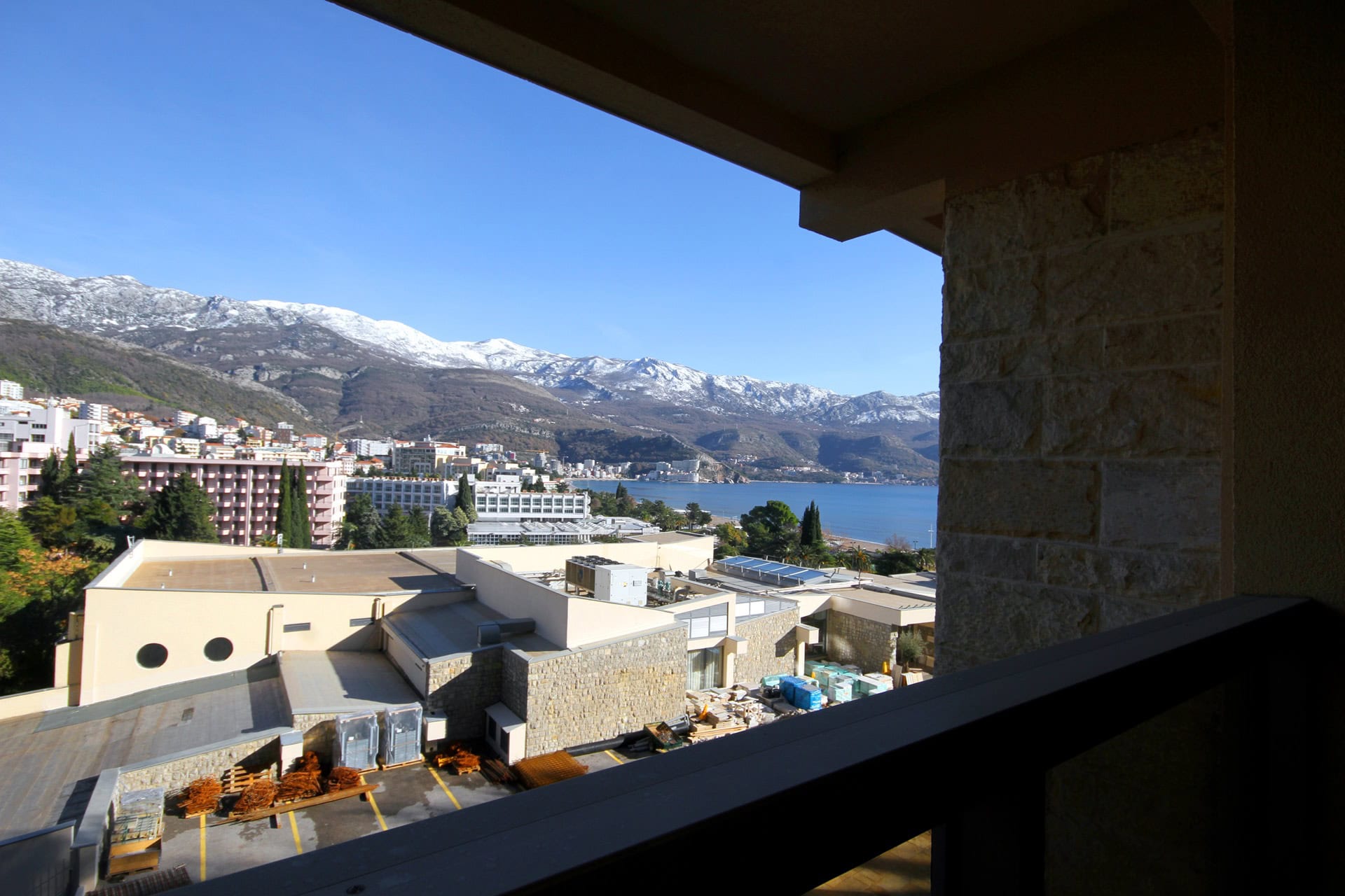 View from the apartment balcony in Budva, Montenegro: a panorama of the sea, the city, and snow-covered mountains on a sunny day.