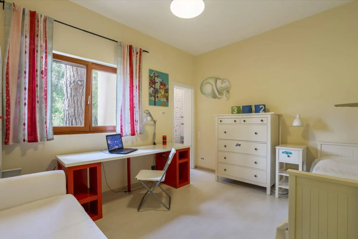 Bright bedroom with a white bed, red study desk holding a laptop, and a white dresser against yellow walls.