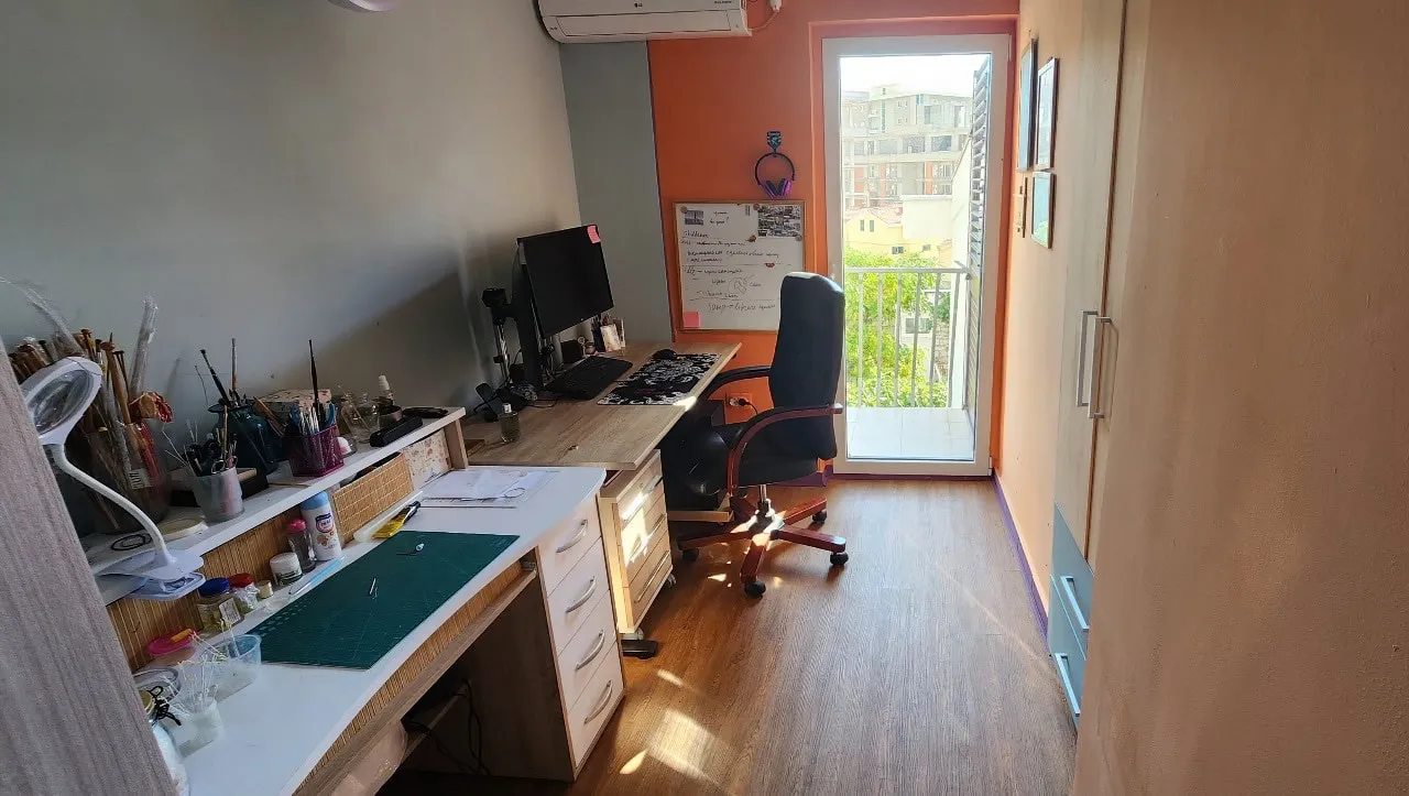 Home office with computer desk, art supplies, wardrobe, and a glass door leading to a balcony.