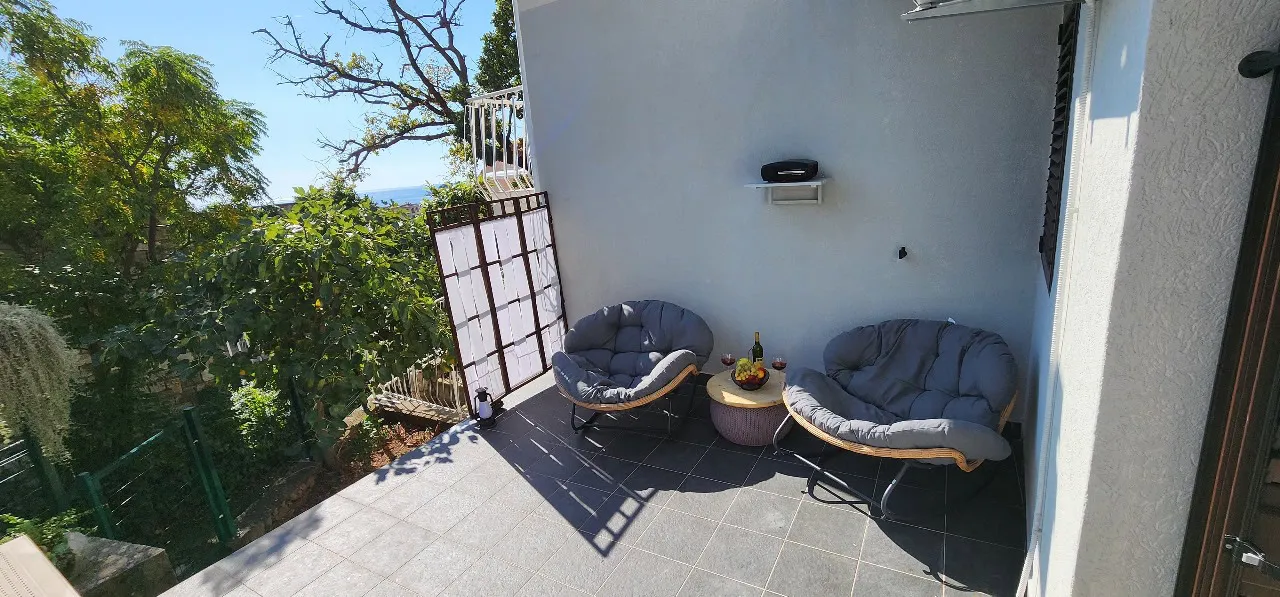 Outdoor terrace with two grey moon chairs and a small table overlooking green trees and the sea.