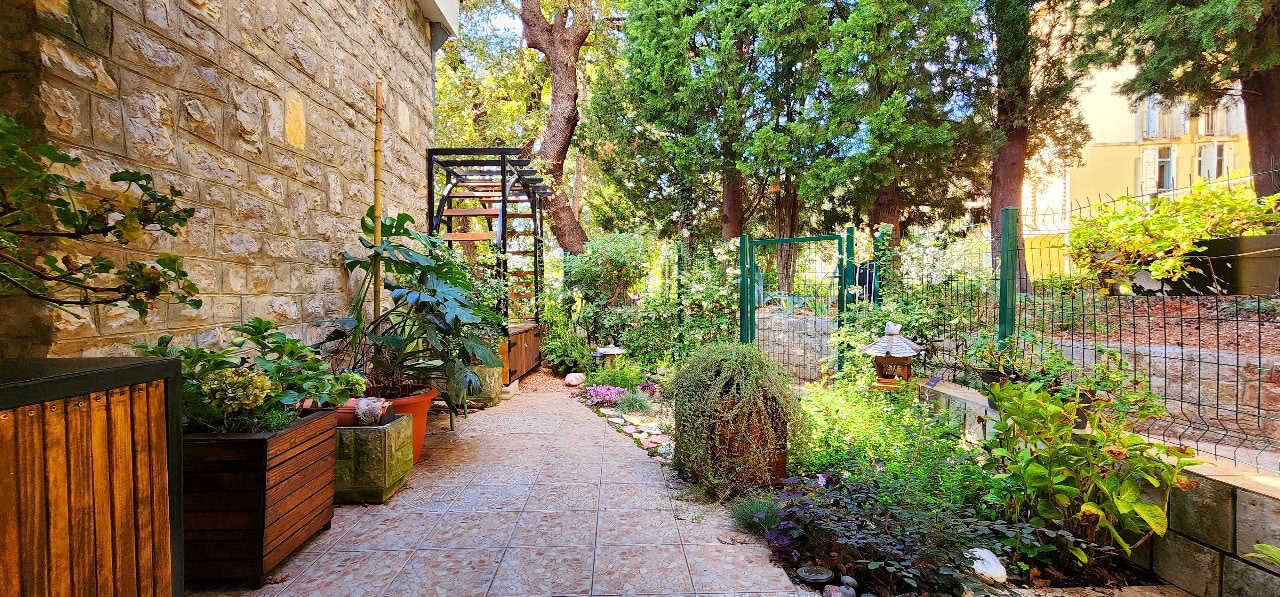 Garden with stone wall, tiled path, potted plants, wooden archway, and trees in the background.