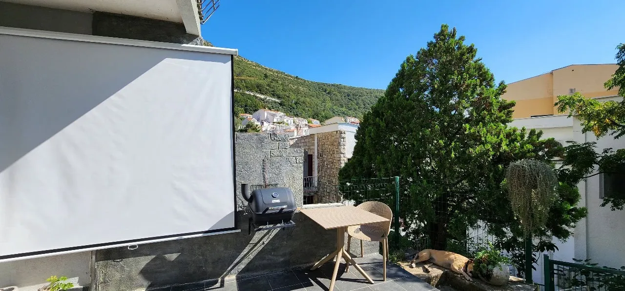 Terrace with projection screen, grill, table and chair overlooking a green hill and blue sky.