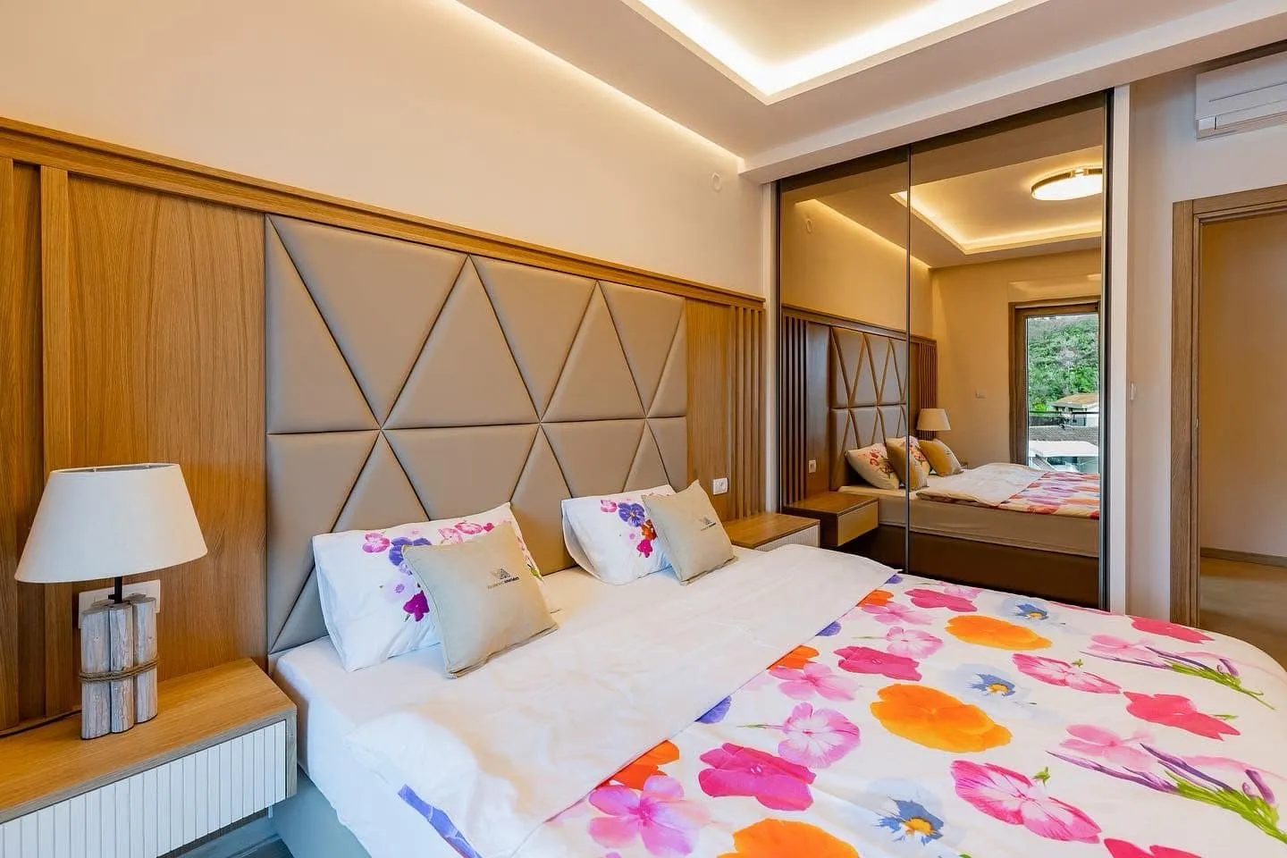 Modern bedroom featuring a large bed with floral bedding, padded headboard, and mirrored wardrobe.