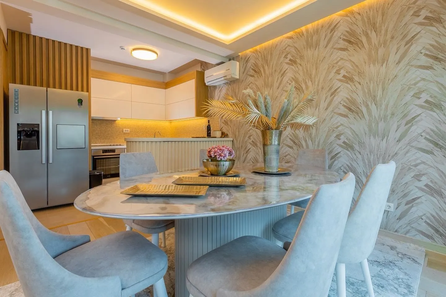 Modern kitchen with a marble dining table, grey chairs, and visible cooking area with a refrigerator.