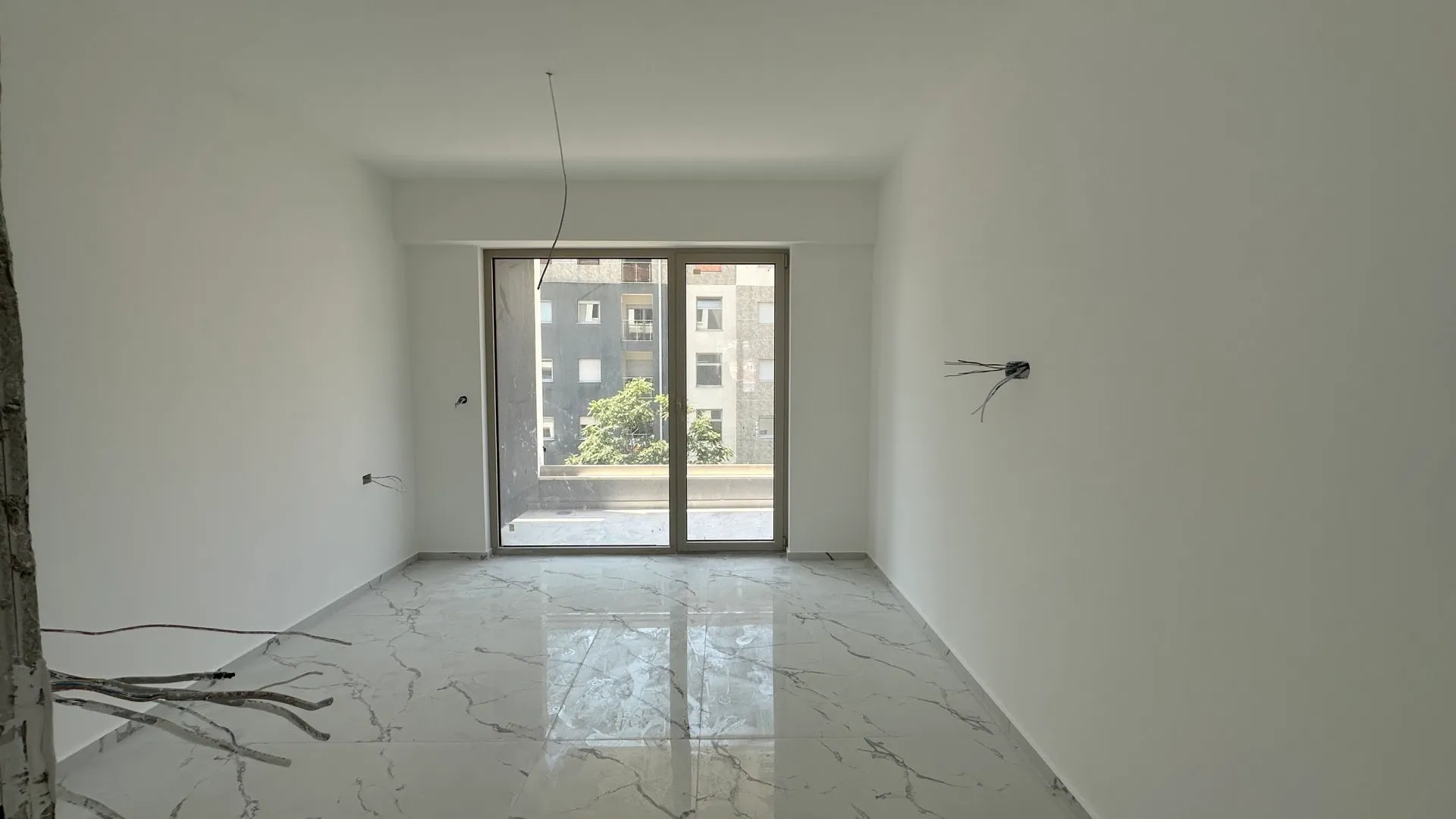Empty room with marble tiles and balcony door, under renovation with exposed wires.
