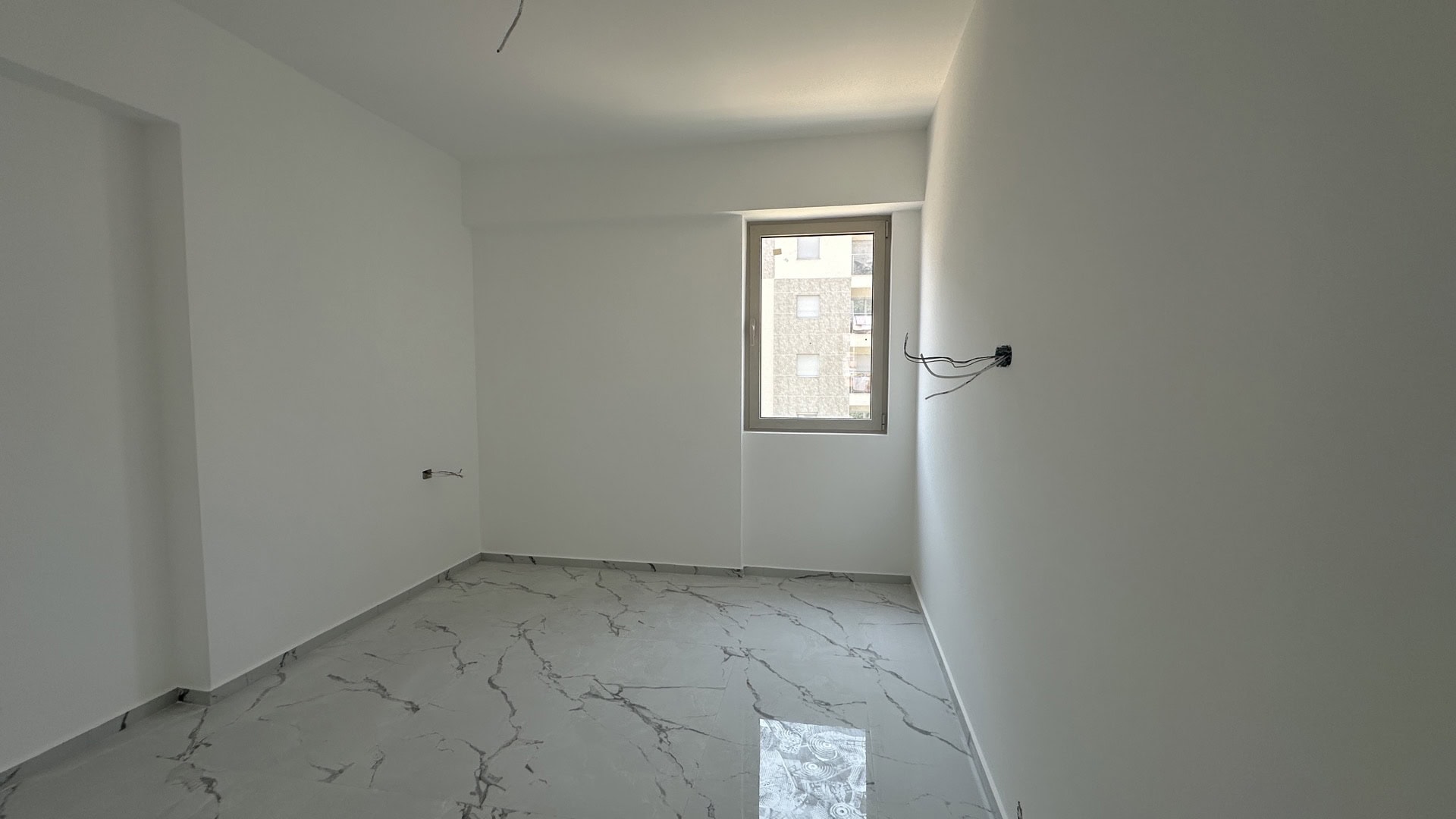 Bright bedroom in a new building in Budva, Montenegro: white walls, marble-look porcelain tile flooring, view from the window.