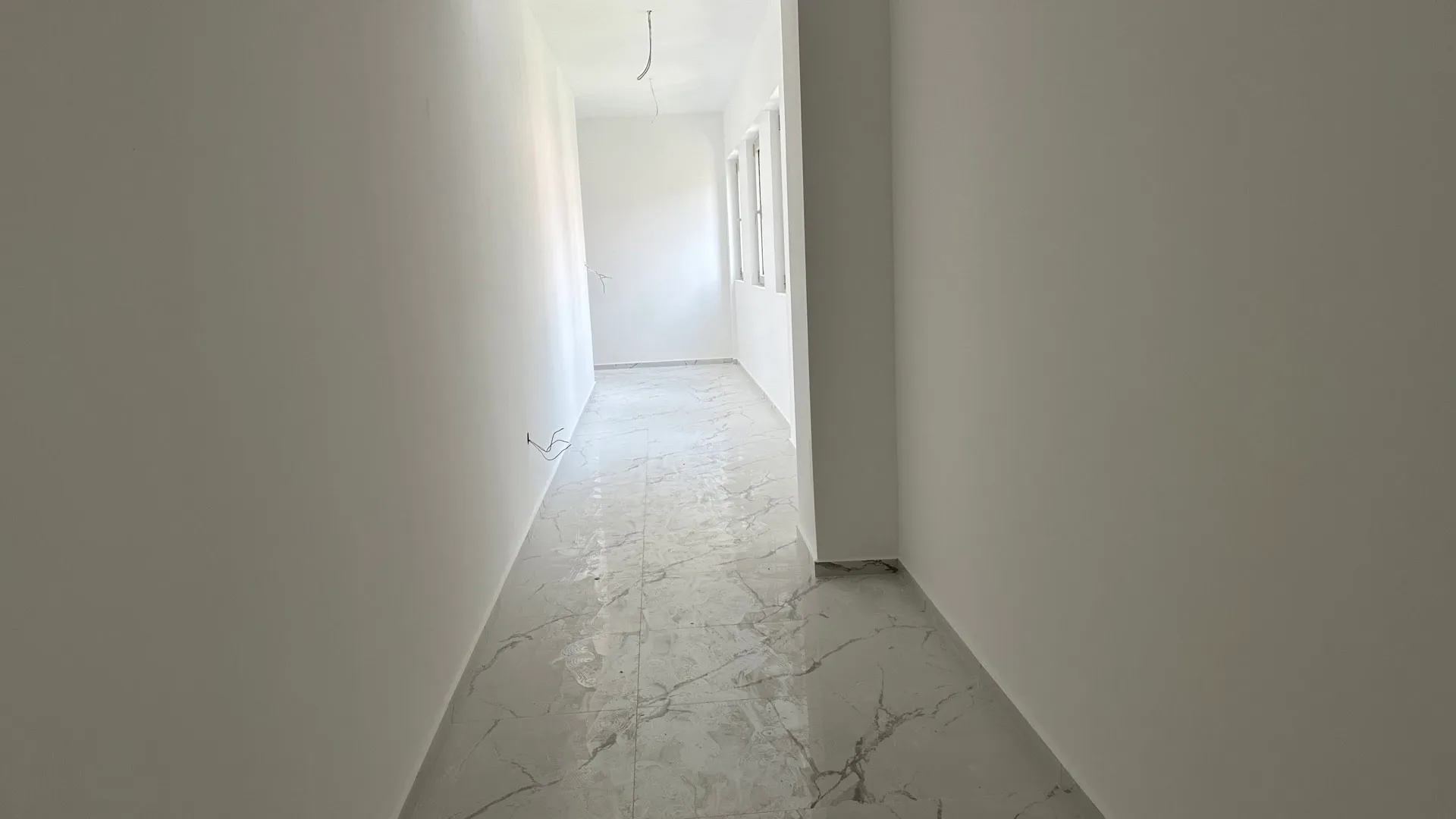 Empty hallway with white walls and light marble-patterned tile flooring. Exposed electrical wires and windows visible at the end.
