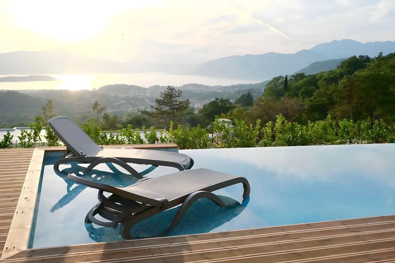 Infinity pool with lounge chairs on a wooden deck overlooking mountains and water at sunset.