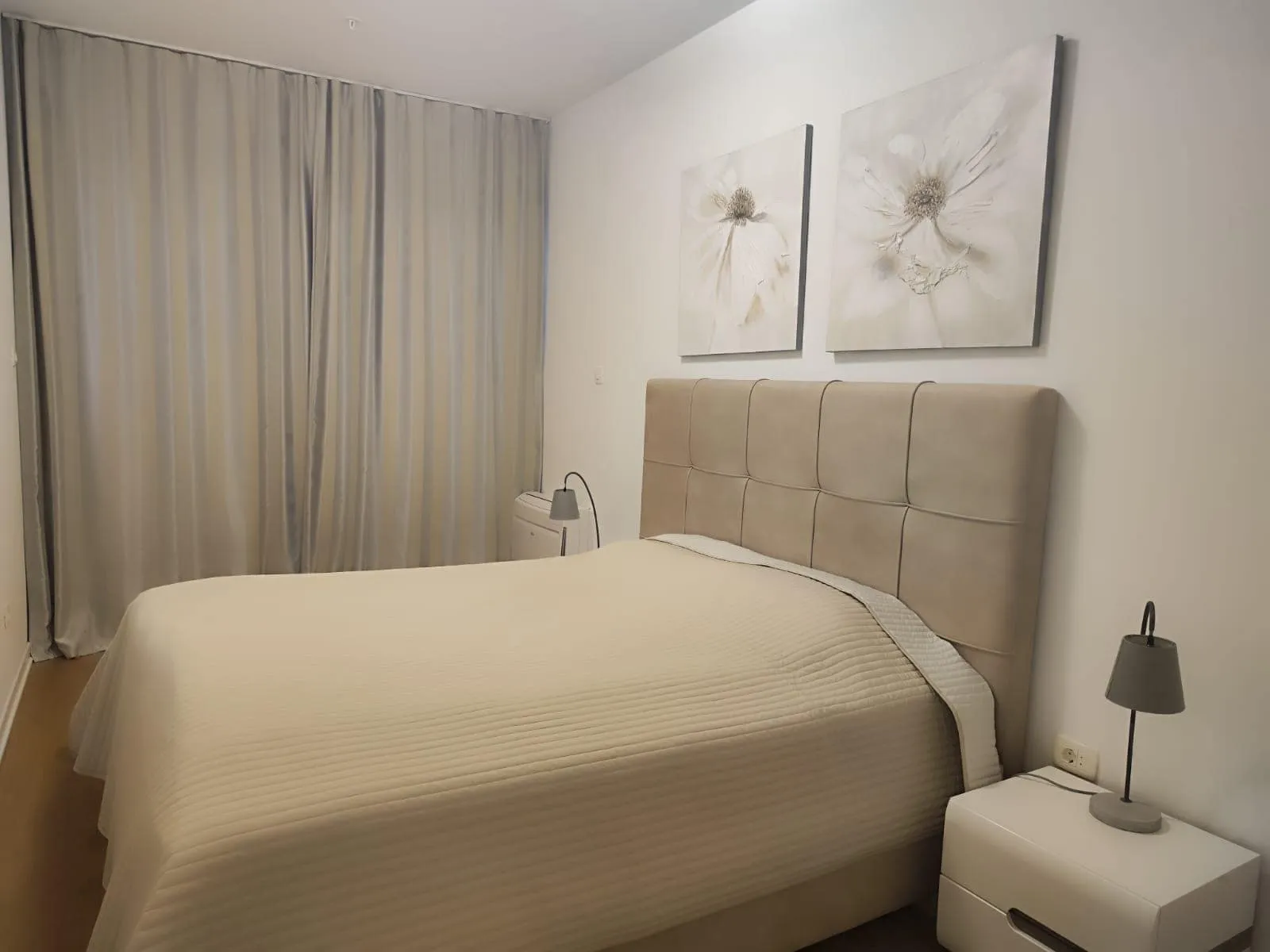 Modern bedroom featuring a double bed with a beige upholstered headboard, floral wall art, and neutral curtains.