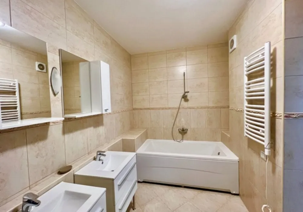 Bathroom with beige tiles, white bathtub, double sink vanity, wall mirror, and electric towel warmer.