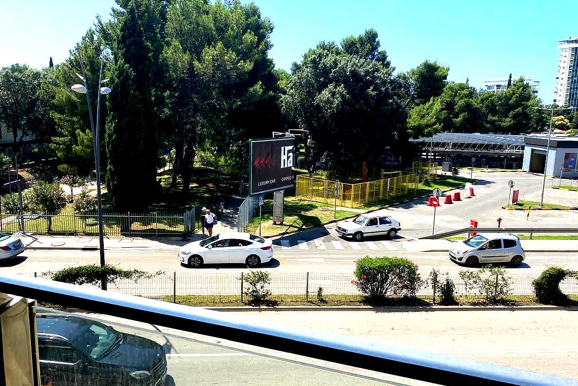 View from the apartment balcony in central Budva, Montenegro: a city street, a green park, and parking next to the building.