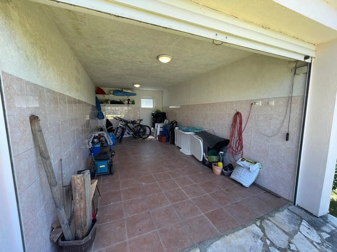 A tiled garage or utility room used for storage, containing bicycles, tools, wood, and various household items under a concrete ceiling.
