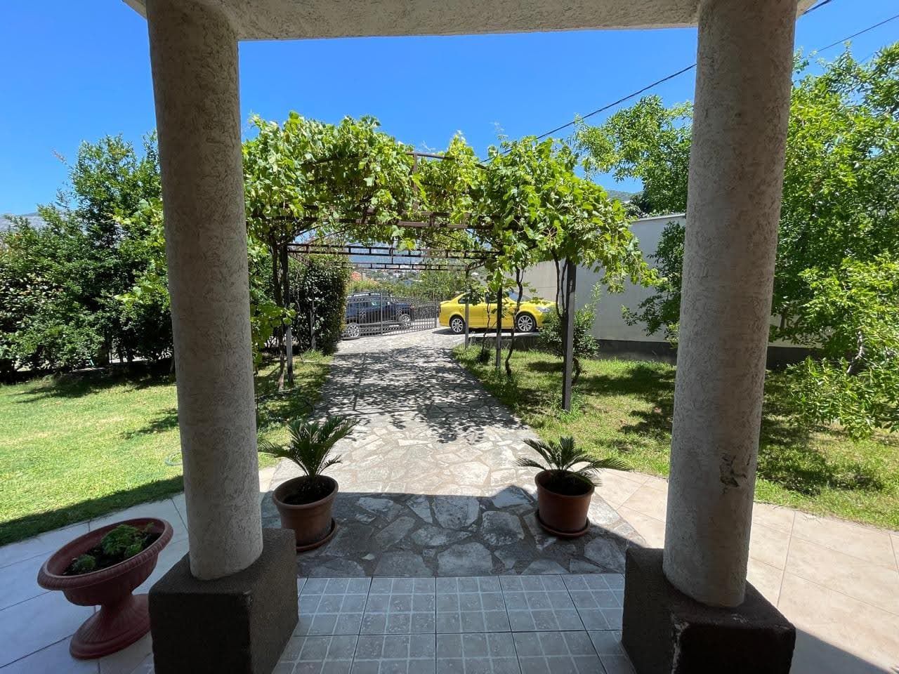 View from the house terrace in Bar, Montenegro: a green garden with a grapevine arch, a stone path, and parking for cars.