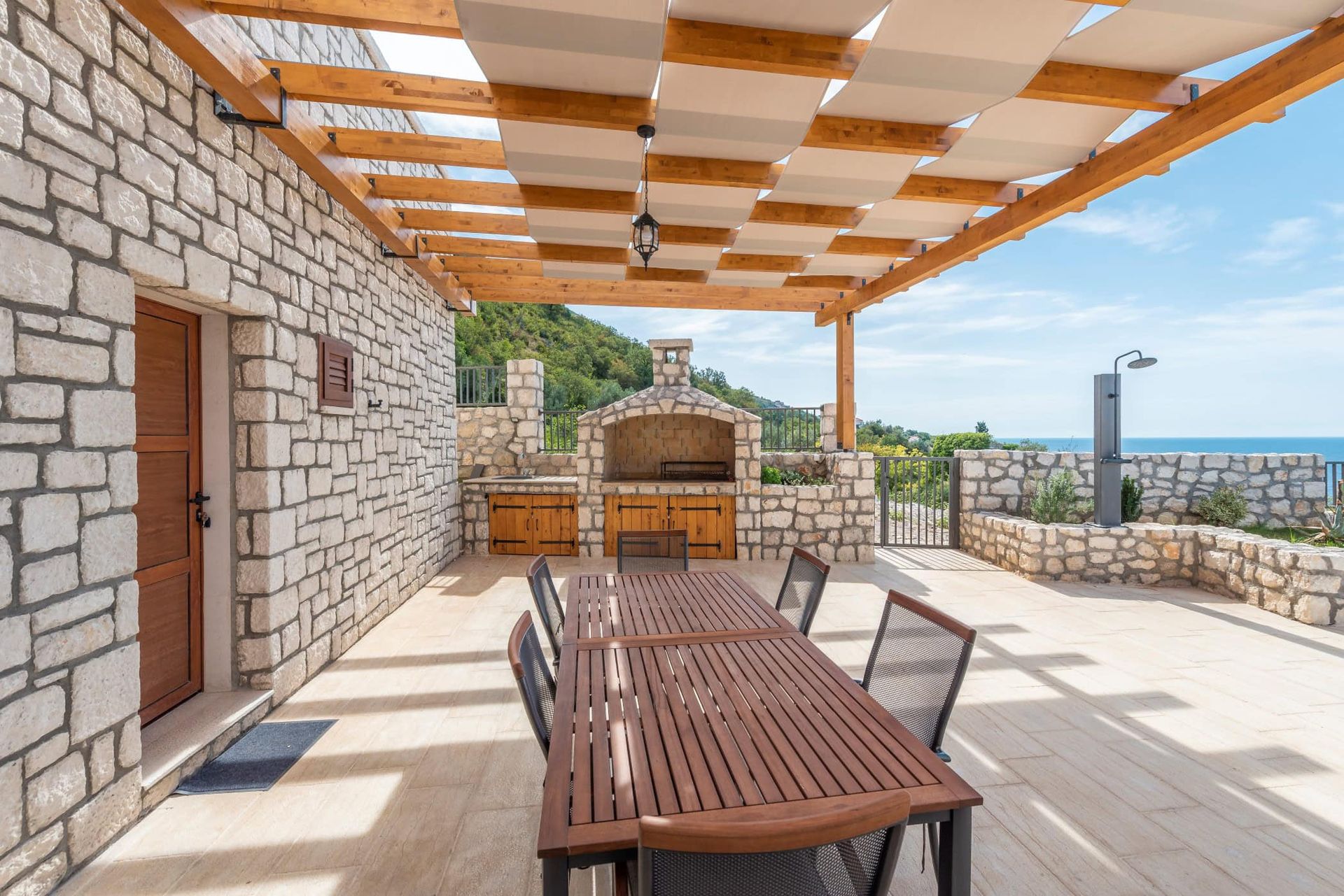 House terrace in Reževići, Montenegro: stone finish, barbecue, wooden table, and sea view under a canopy.