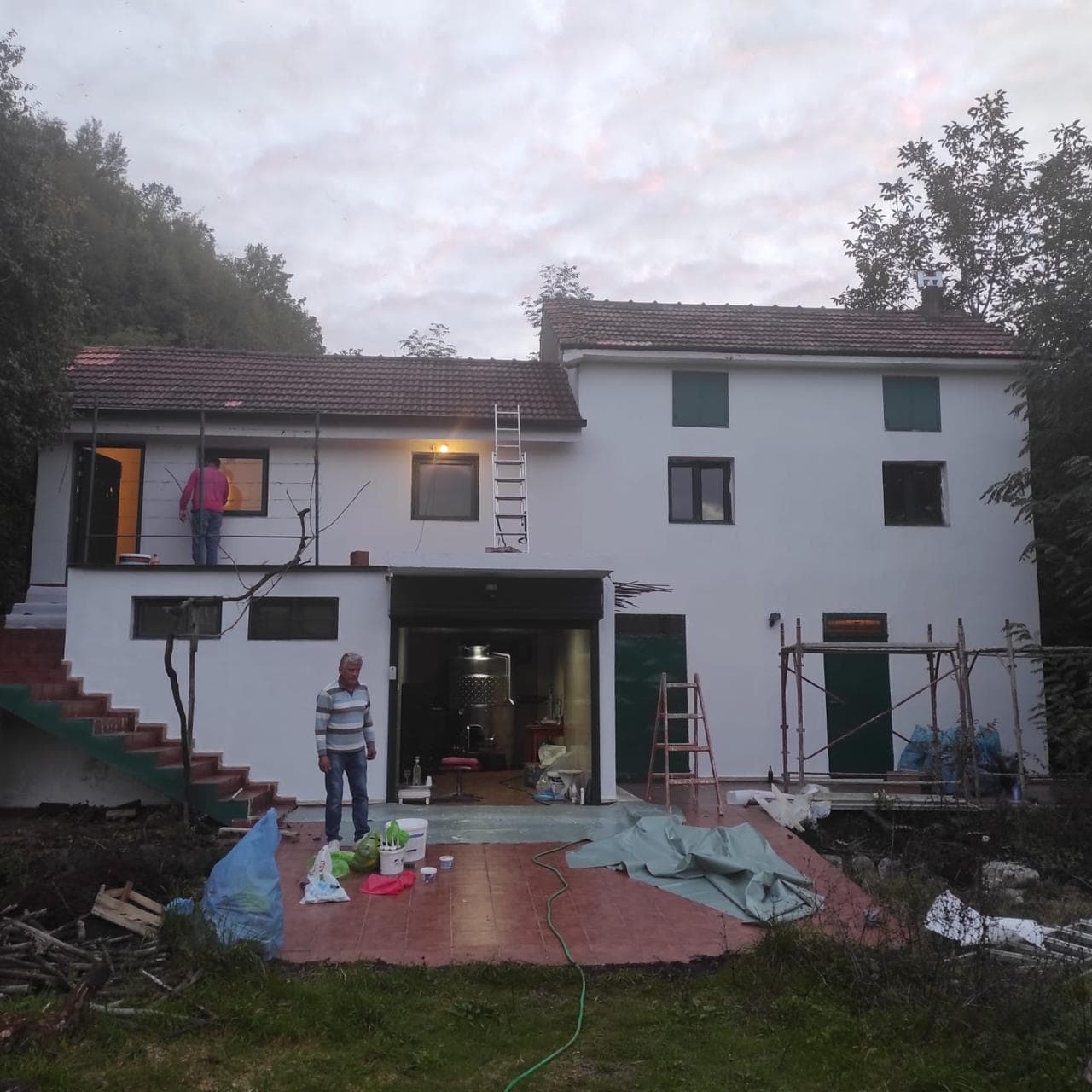 Exterior of a two-story house under renovation with scaffolding, workers, construction materials, and an open garage door.