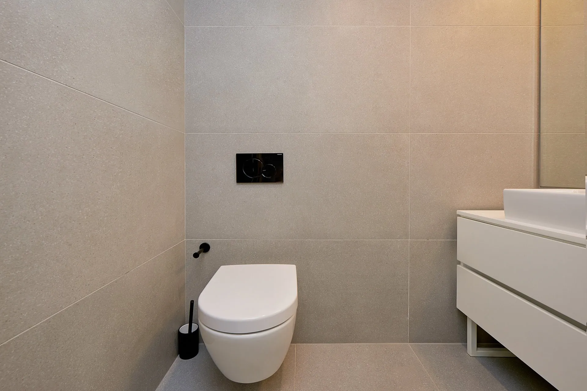 Modern bathroom featuring a wall-mounted toilet with a black flush plate and a white vanity unit with a sink on light tiles.