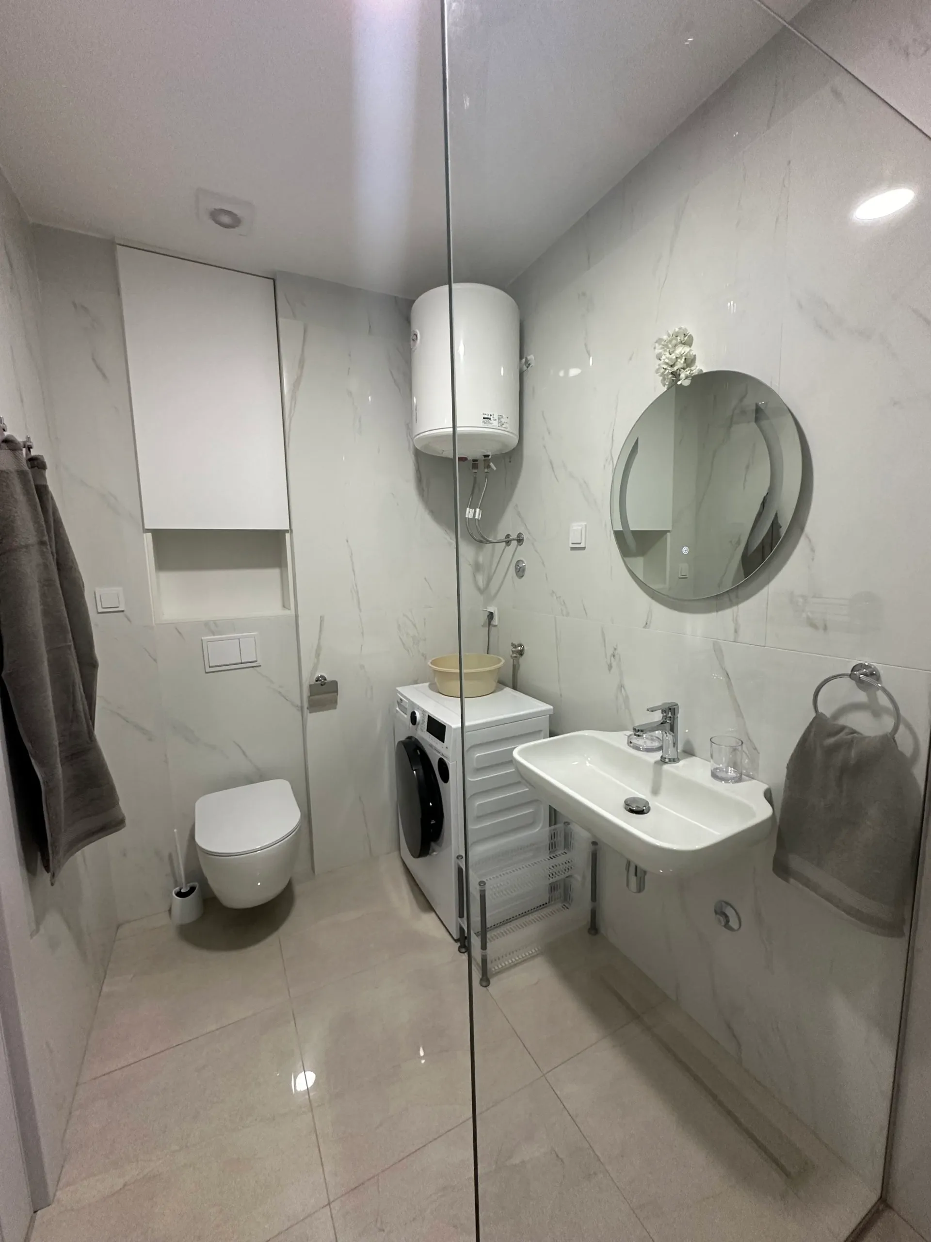 Bathroom featuring a white toilet, sink, round mirror, and washing machine against marble-patterned wall tiles.