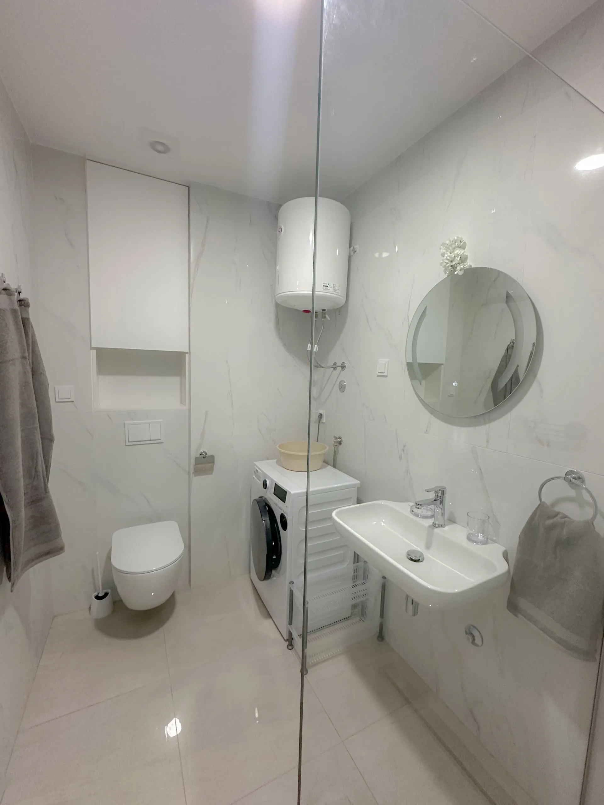 White bathroom featuring a toilet, sink, washing machine, and round mirror on marble-look tiles behind glass.