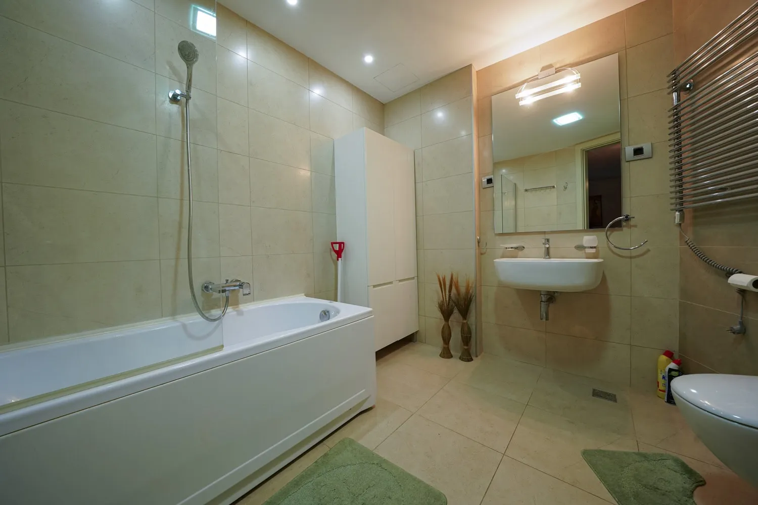Bathroom featuring a white bathtub, shower fixture, wall-mounted toilet, sink with mirror, and beige tiled walls and floor.