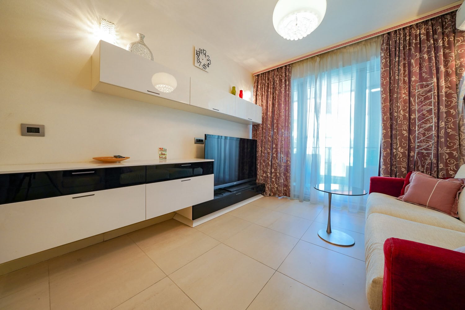 Living room in an apartment in Budva, Montenegro: modern furniture, TV area, light tile flooring, and a panoramic window with curtains.