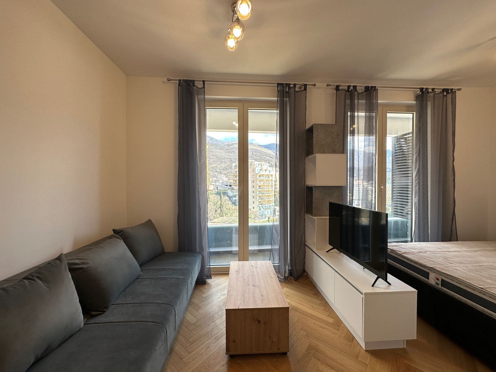 Living room in an apartment in Bečići, Montenegro: gray sofa, TV area, panoramic windows with mountain views, and parquet flooring.