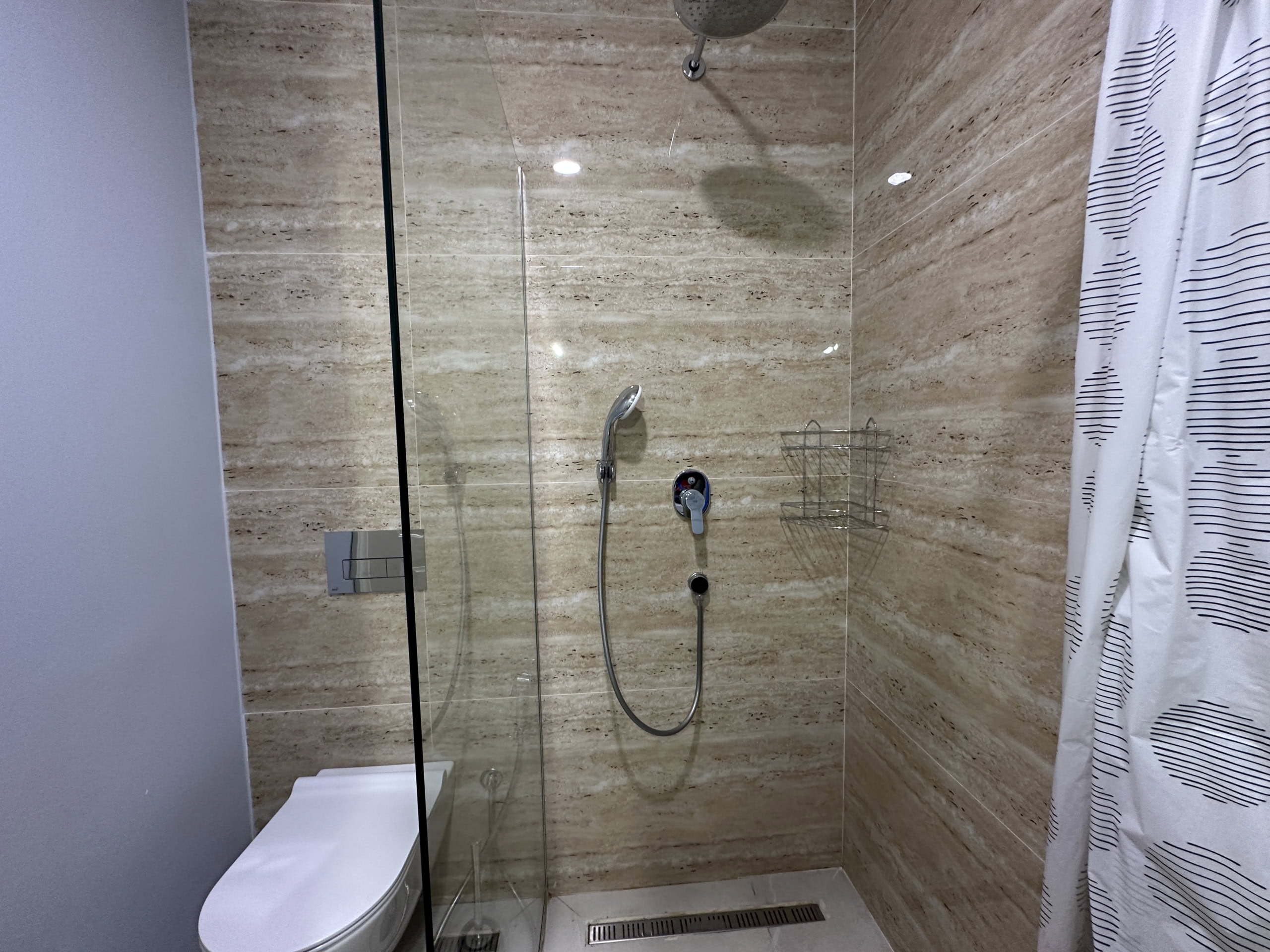 Modern shower area with beige marble-look tile, glass partition, and tropical rain shower in an apartment in Bečići, Montenegro...