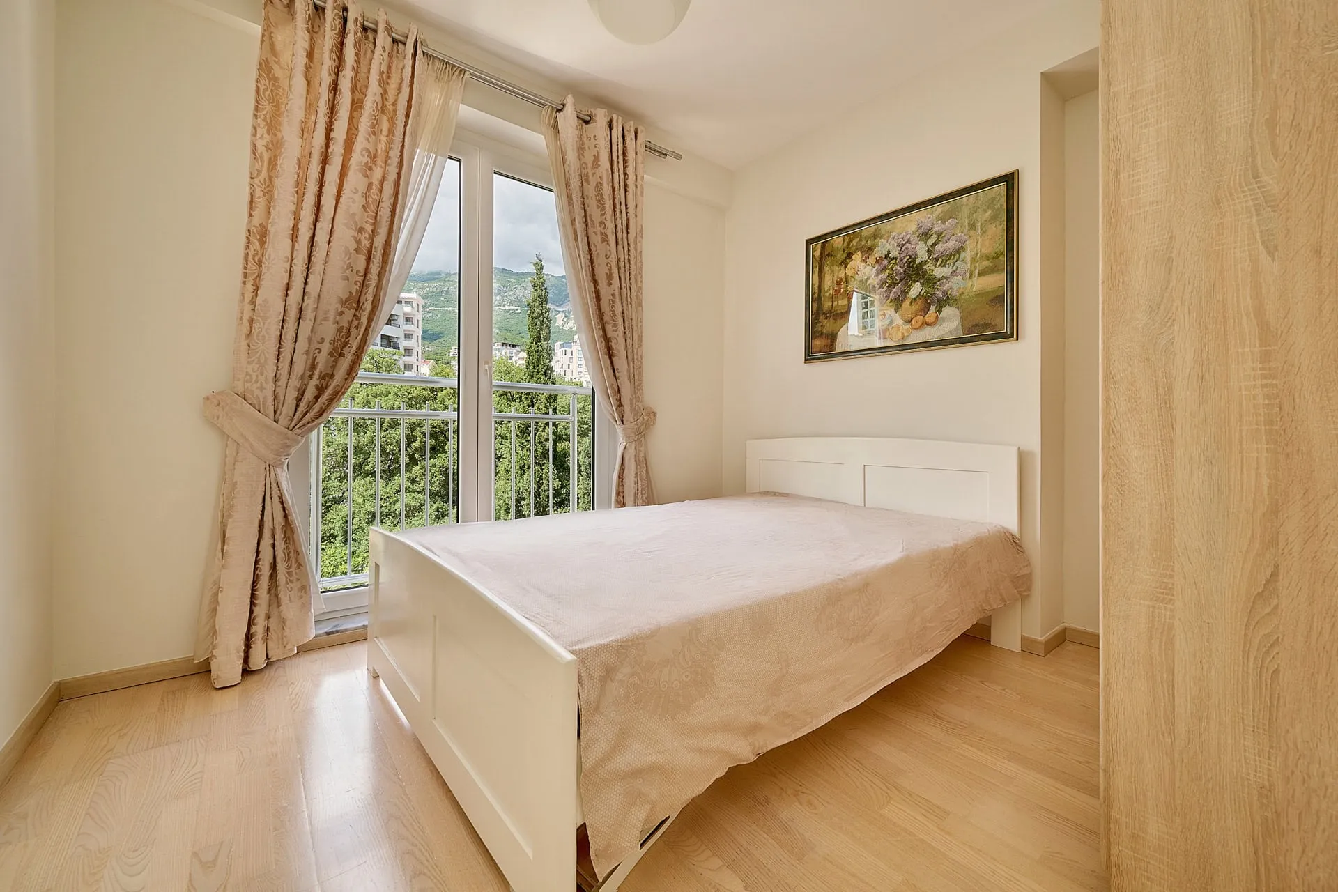 Bright bedroom with a white bed, beige curtains, and a floral painting. A window overlooks a balcony and greenery.