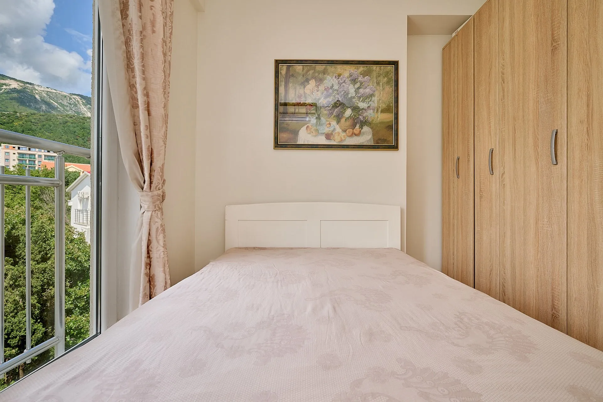 Bedroom featuring a single bed, wooden wardrobe, and floral painting above the headboard near a window.