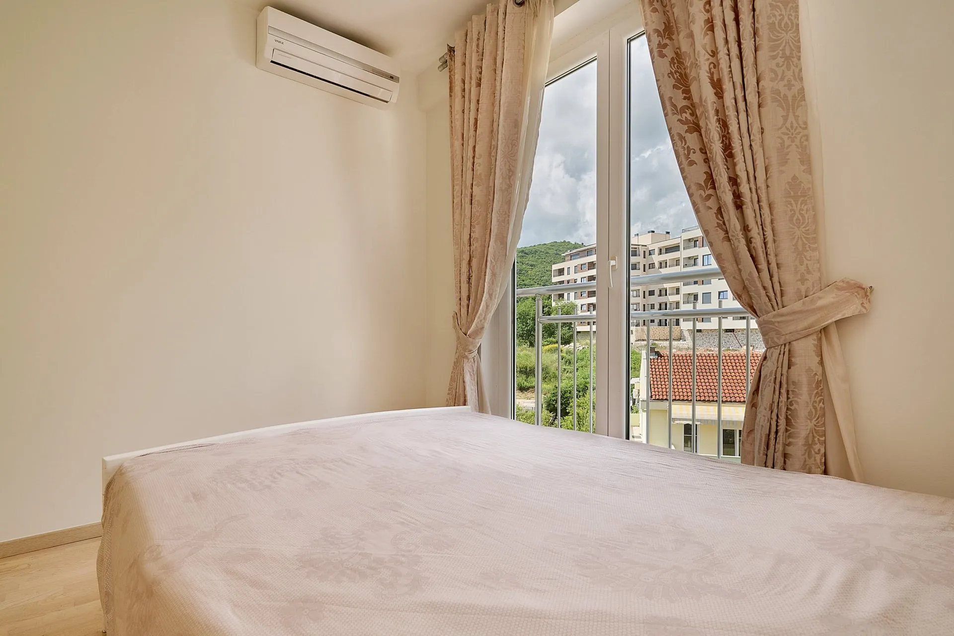Bedroom featuring a bed with light bedding, wall-mounted AC unit, and glass doors opening to a balcony with building views.