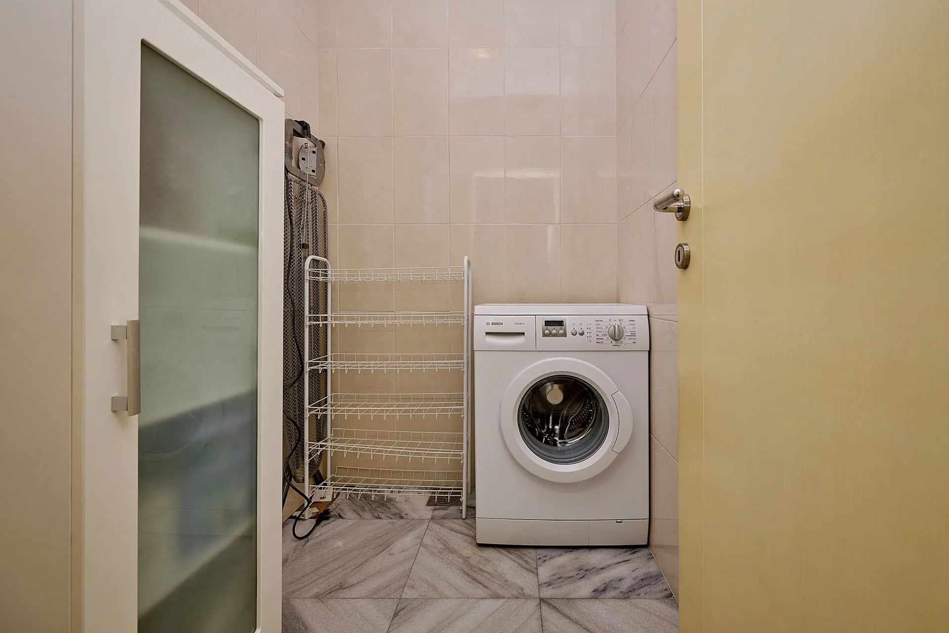 Small utility room featuring a white washing machine, wire shelving unit, and iron against beige tiled walls.