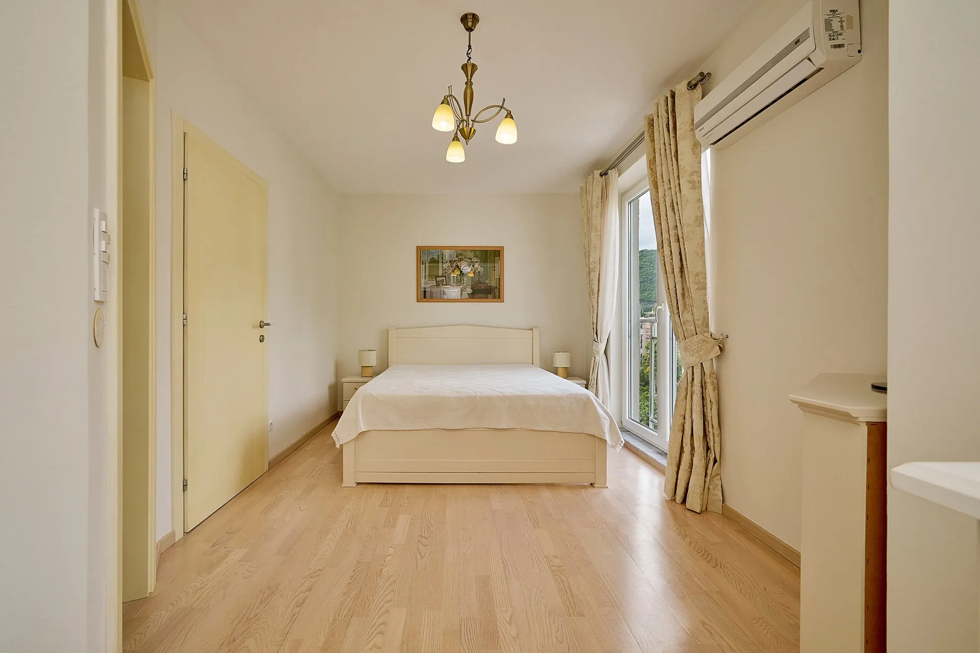 Bright bedroom with wood floors, white bed, chandelier, and wall-mounted AC unit near window.