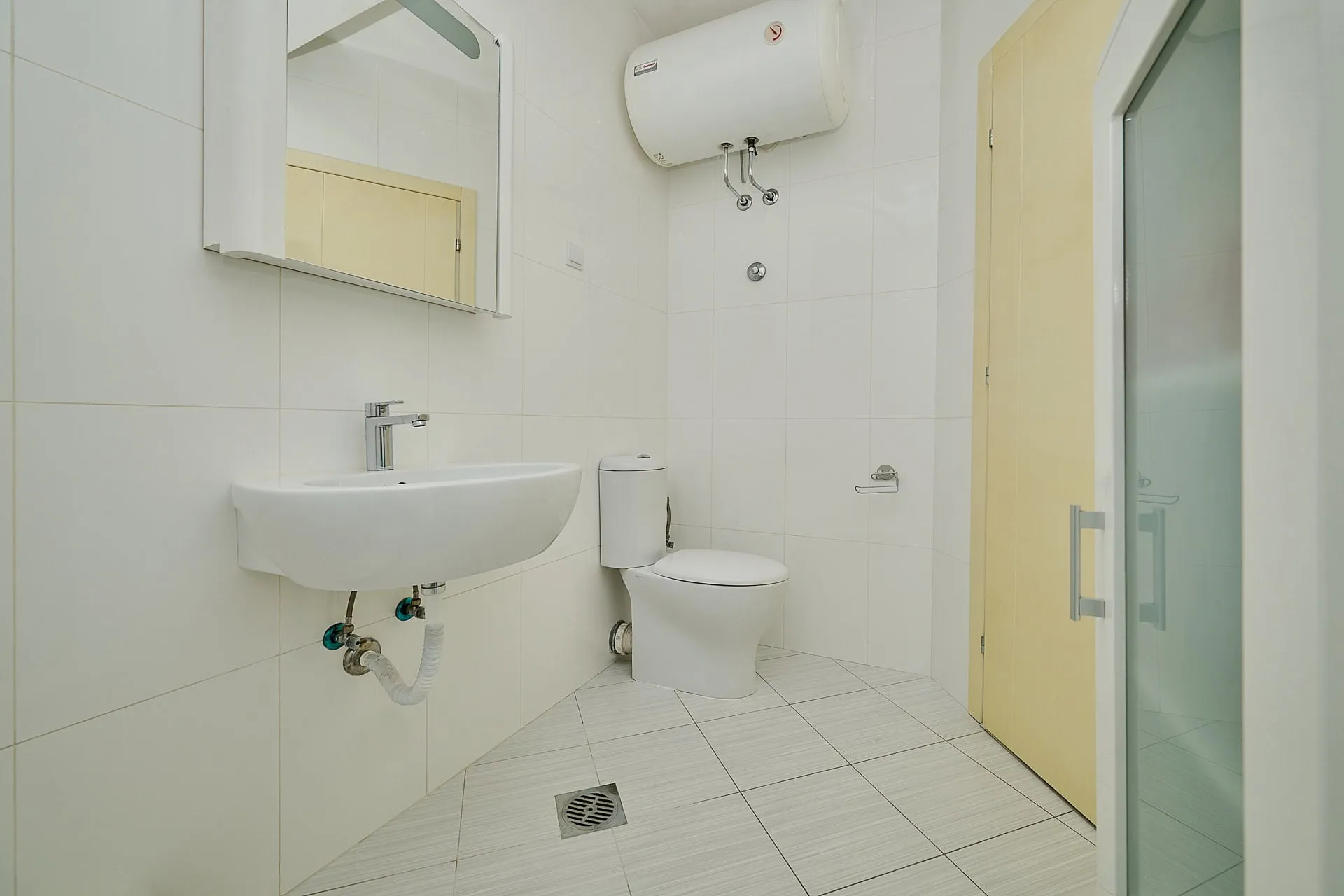 Bathroom with white toilet, wall-mounted sink, mirror, and water heater on tiled walls next to a glass door.