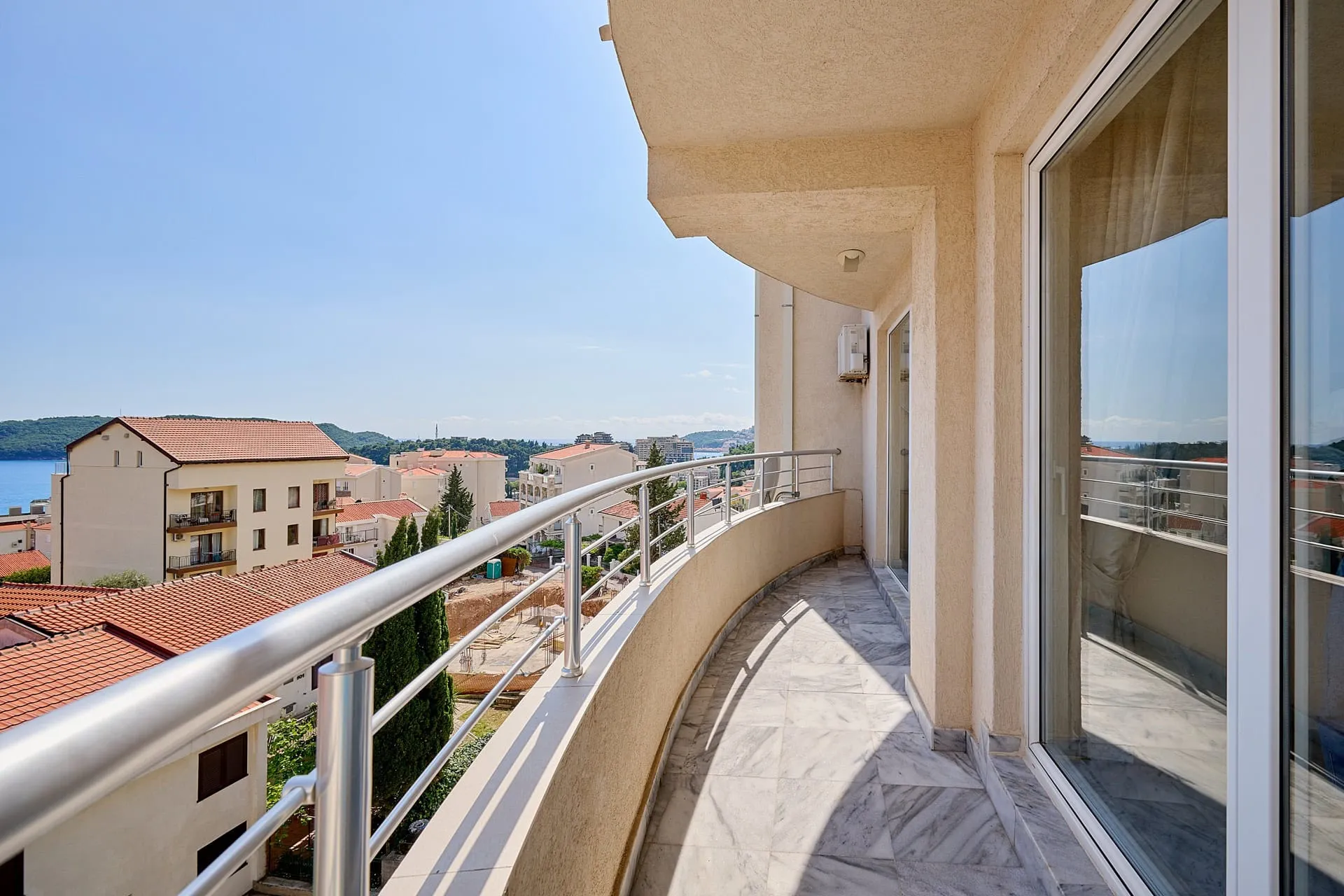 Long curved balcony with marble tiles and metal railing overlooking a coastal town and the sea.