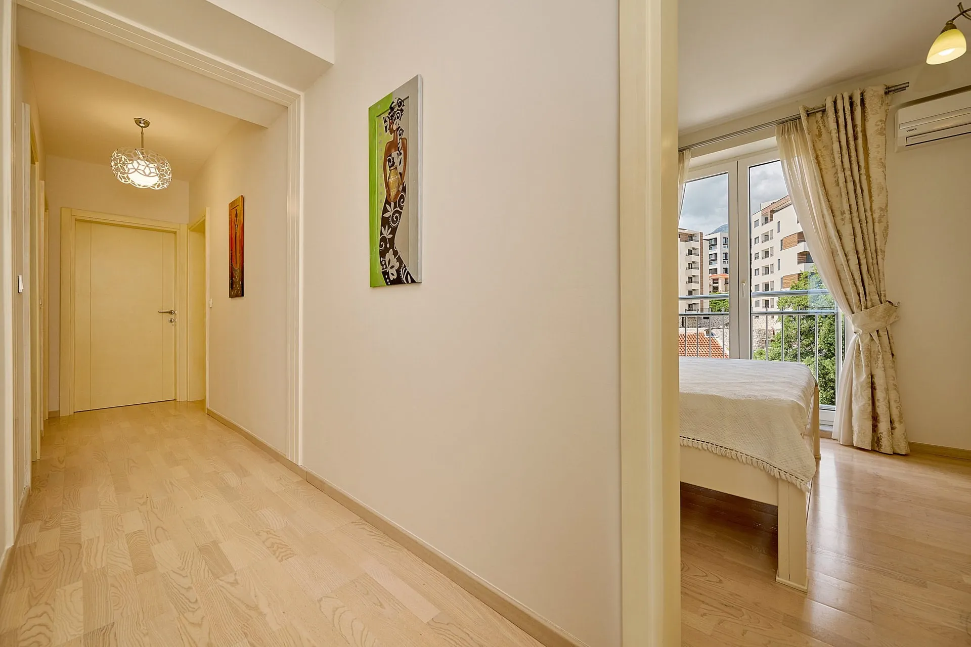 Hallway with light wood flooring and artwork, partial view of a bedroom with a bed by the window.