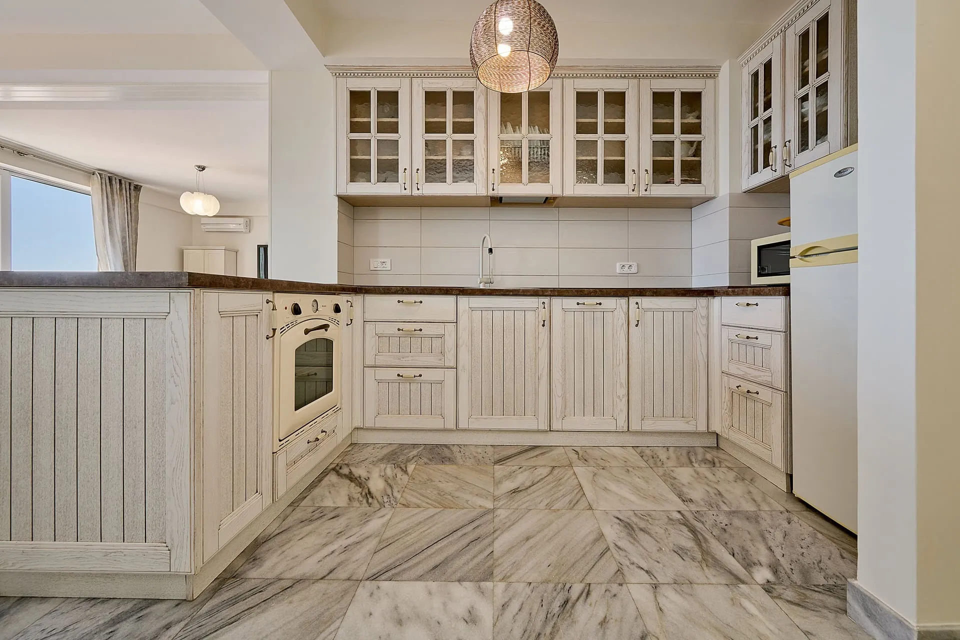 Bright kitchen with white wood cabinets, marble floor, built-in oven, refrigerator, and wicker pendant light.