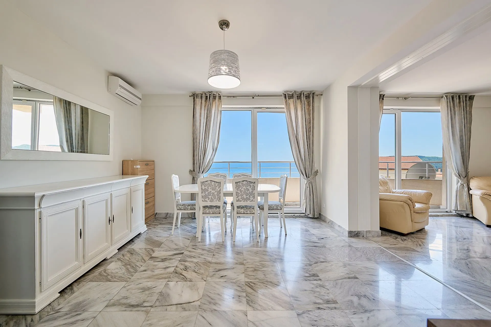 Bright room with marble floor, white dining set, beige sofa, and large windows overlooking the sea.