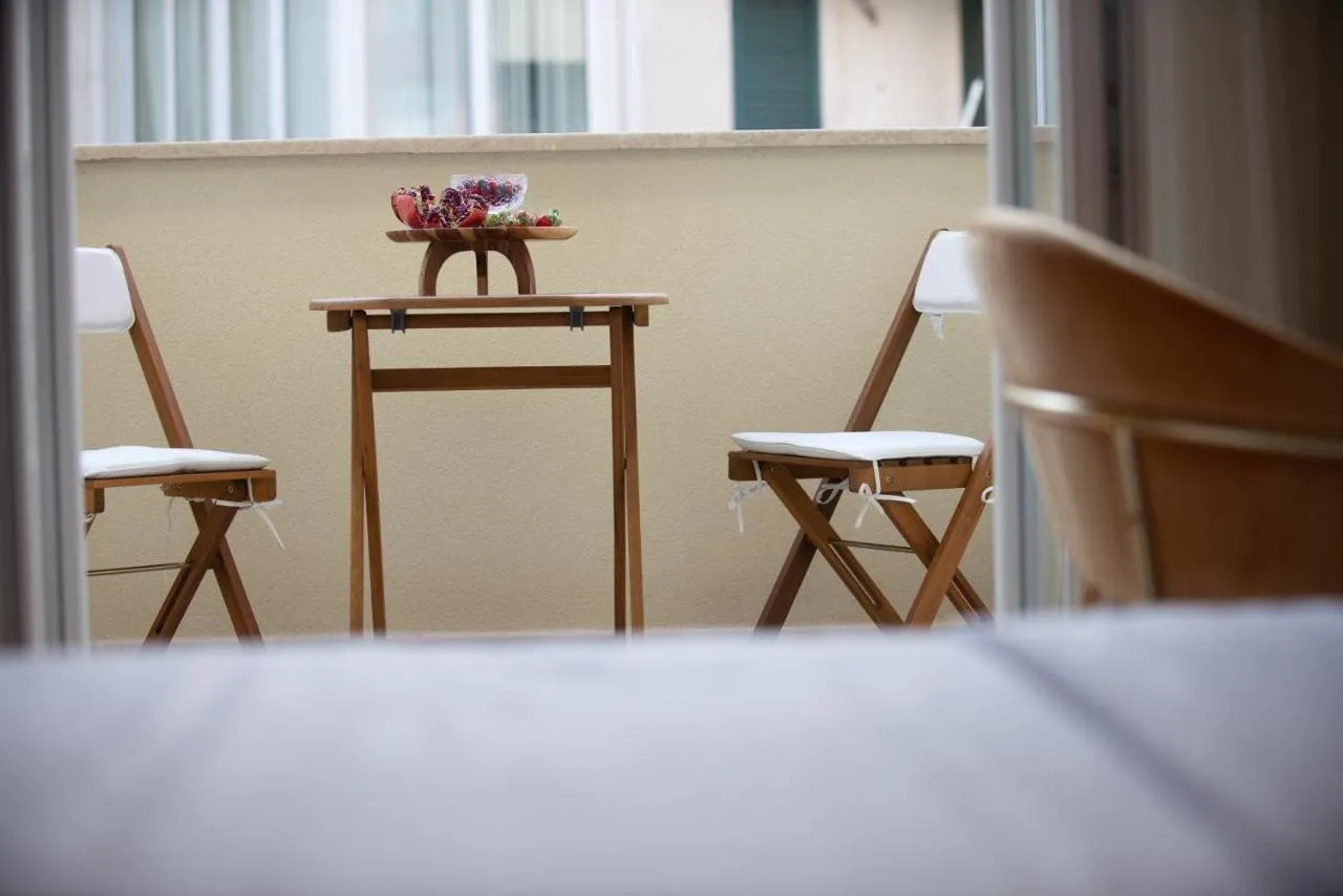 Wooden table and two folding chairs with white cushions on a balcony facing a beige wall.