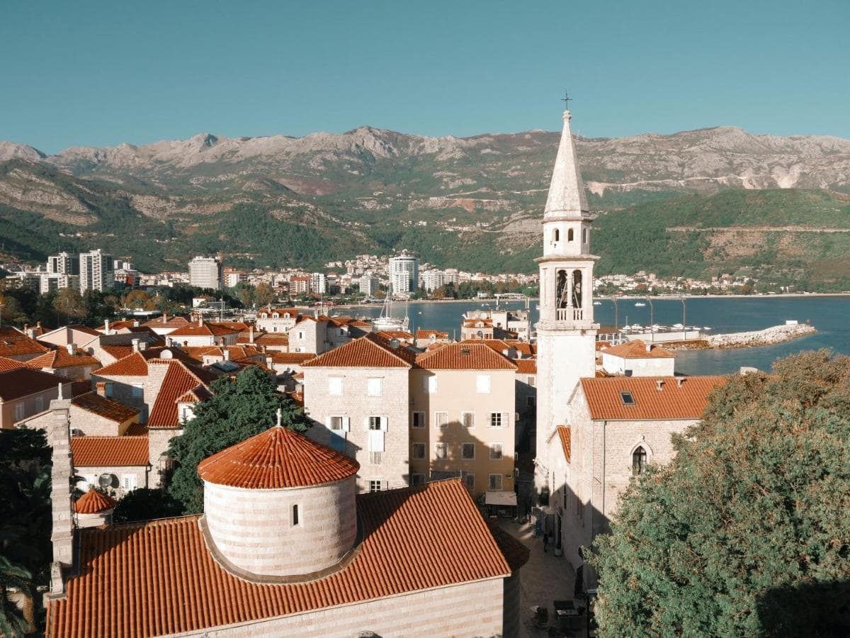 Panoramic view of the old town of Budva, Montenegro: tiled roofs, a church, and the sea bay against a mountain backdrop.