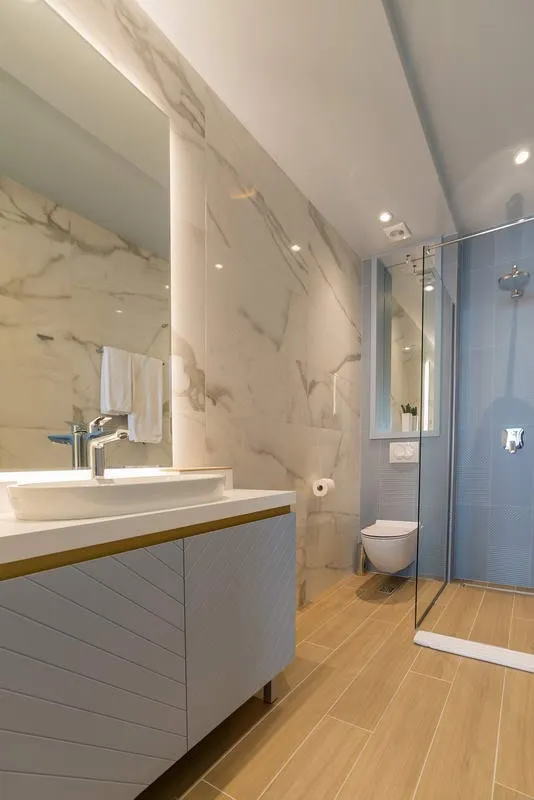 Modern bathroom featuring marble walls, a vessel sink on a vanity, wall-mounted toilet, and a glass-enclosed shower with blue tiles.
