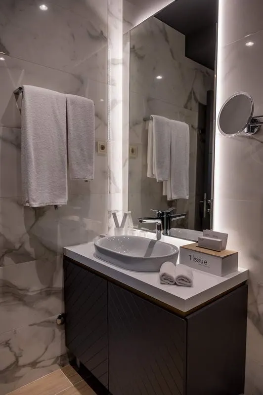 Modern bathroom with marble walls, vessel sink, large illuminated mirror, and white towels on a rack.