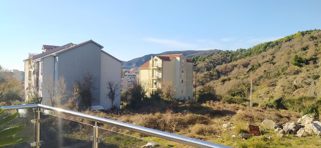 View from the balcony of an apartment in Bečići, Montenegro: modern residential buildings, mountain landscape, and green slopes against the sky.
