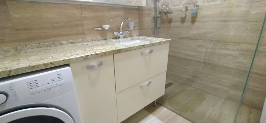 Bathroom with sink, vanity cabinet, washing machine, and glass shower partition against beige tiled walls.