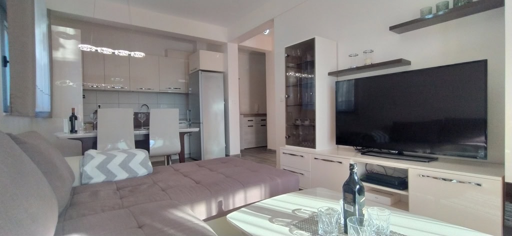 Modern living room with kitchen in an apartment in Bečići, Montenegro: corner sofa, TV area, and dining furniture.